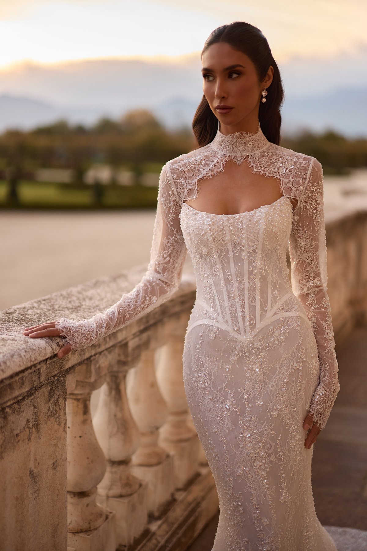 Woman in elegant white beaded wedding dress with long lace sleeves standing by stone railing at sunset
