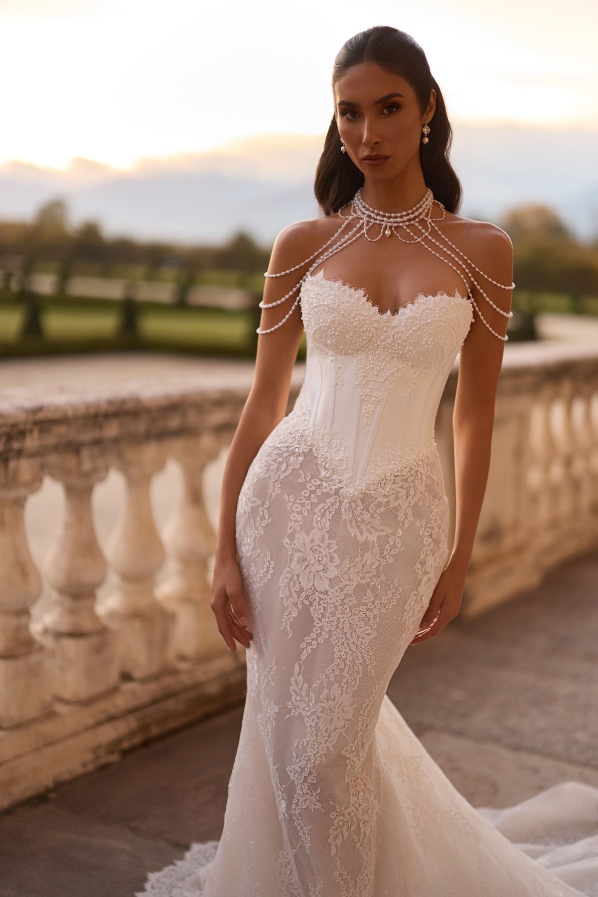 Woman in white lace wedding dress with beaded shoulder jewelry posing on terrace at sunset with mountains in background