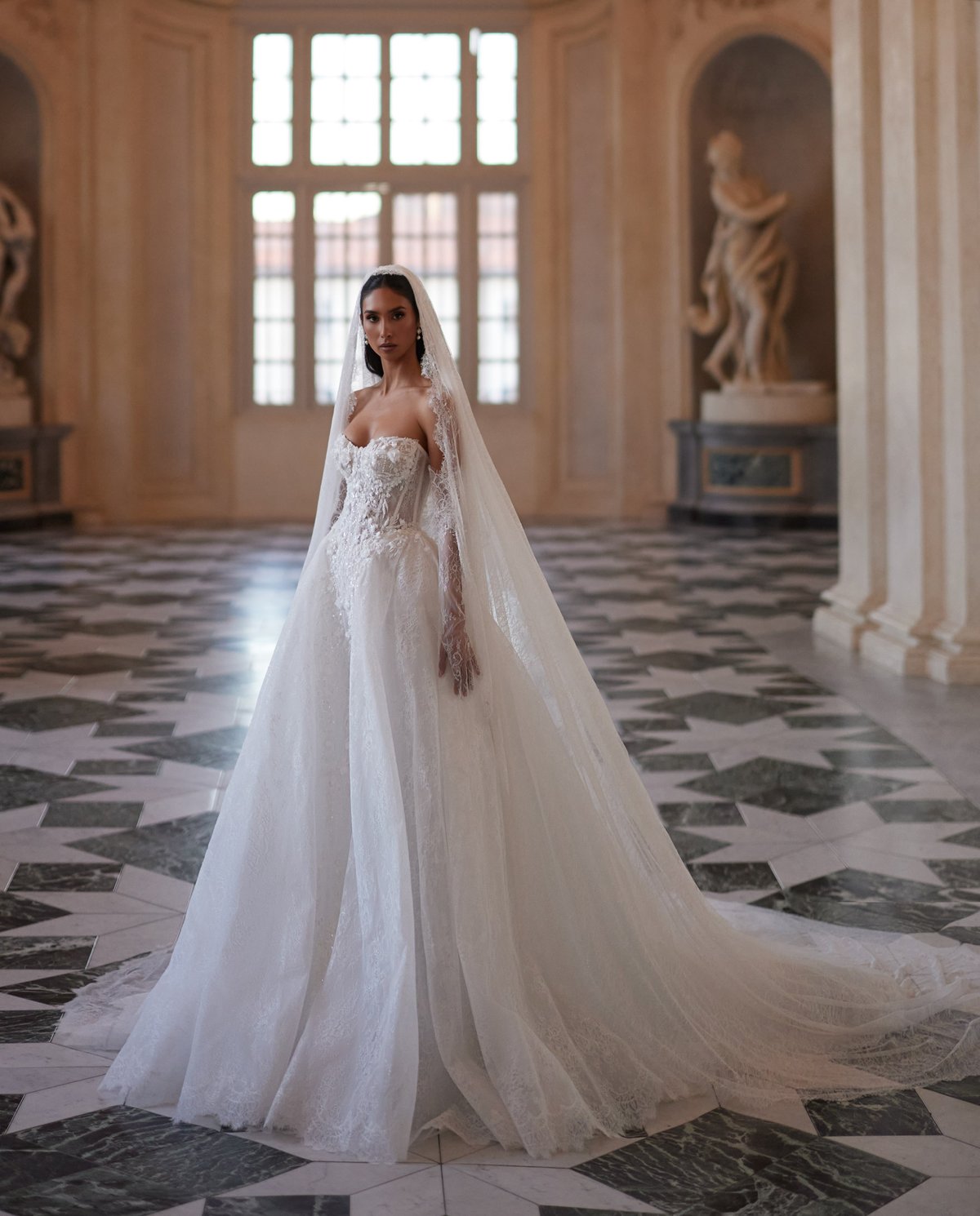 Bride in white wedding gown with veil and train standing in ornate hall with patterned floor and classical sculptures
