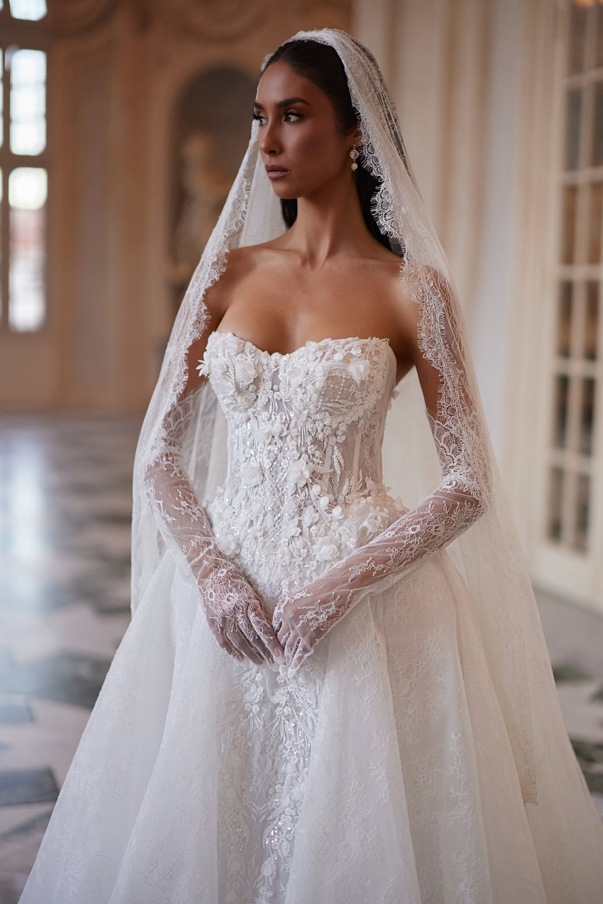 Bride in elegant white strapless wedding dress with beaded bodice and lace veil, standing in ornate hallway