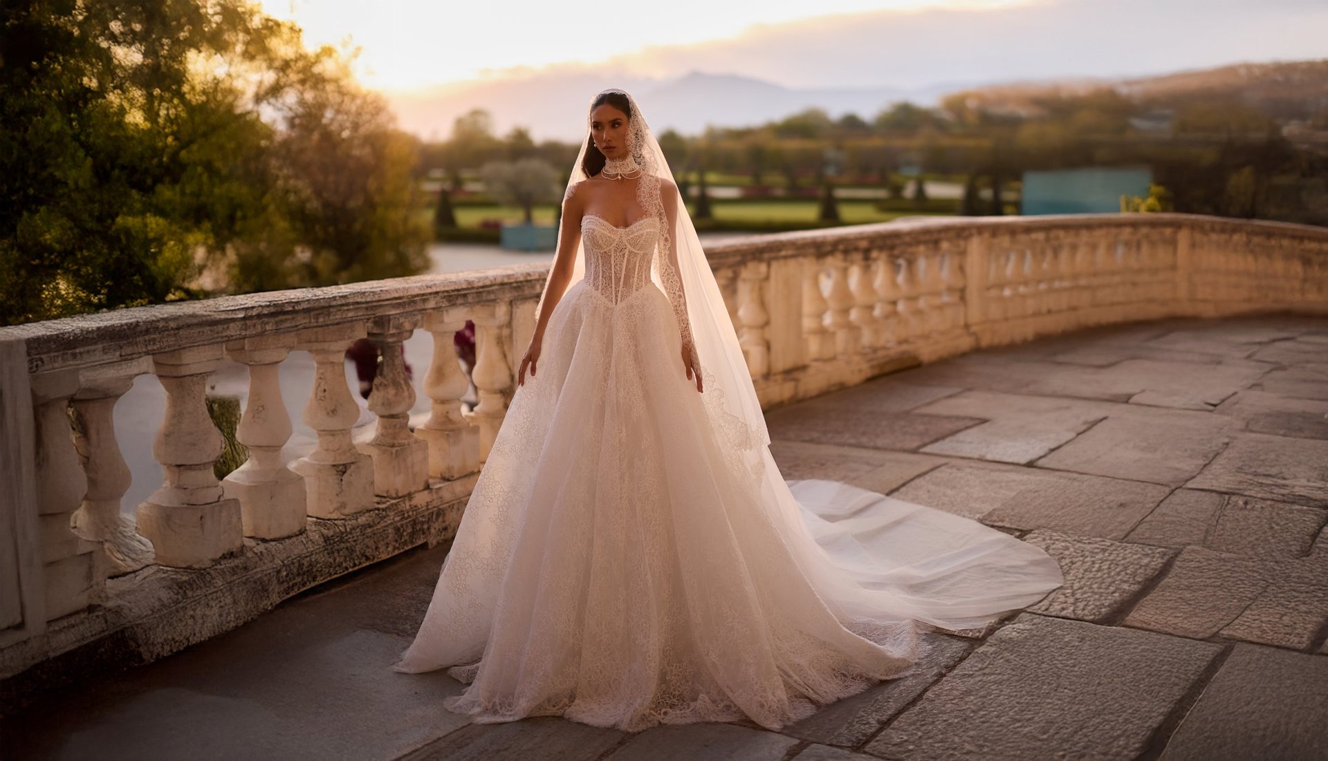 Bride in white wedding gown with veil standing on stone terrace overlooking scenic landscape at sunset