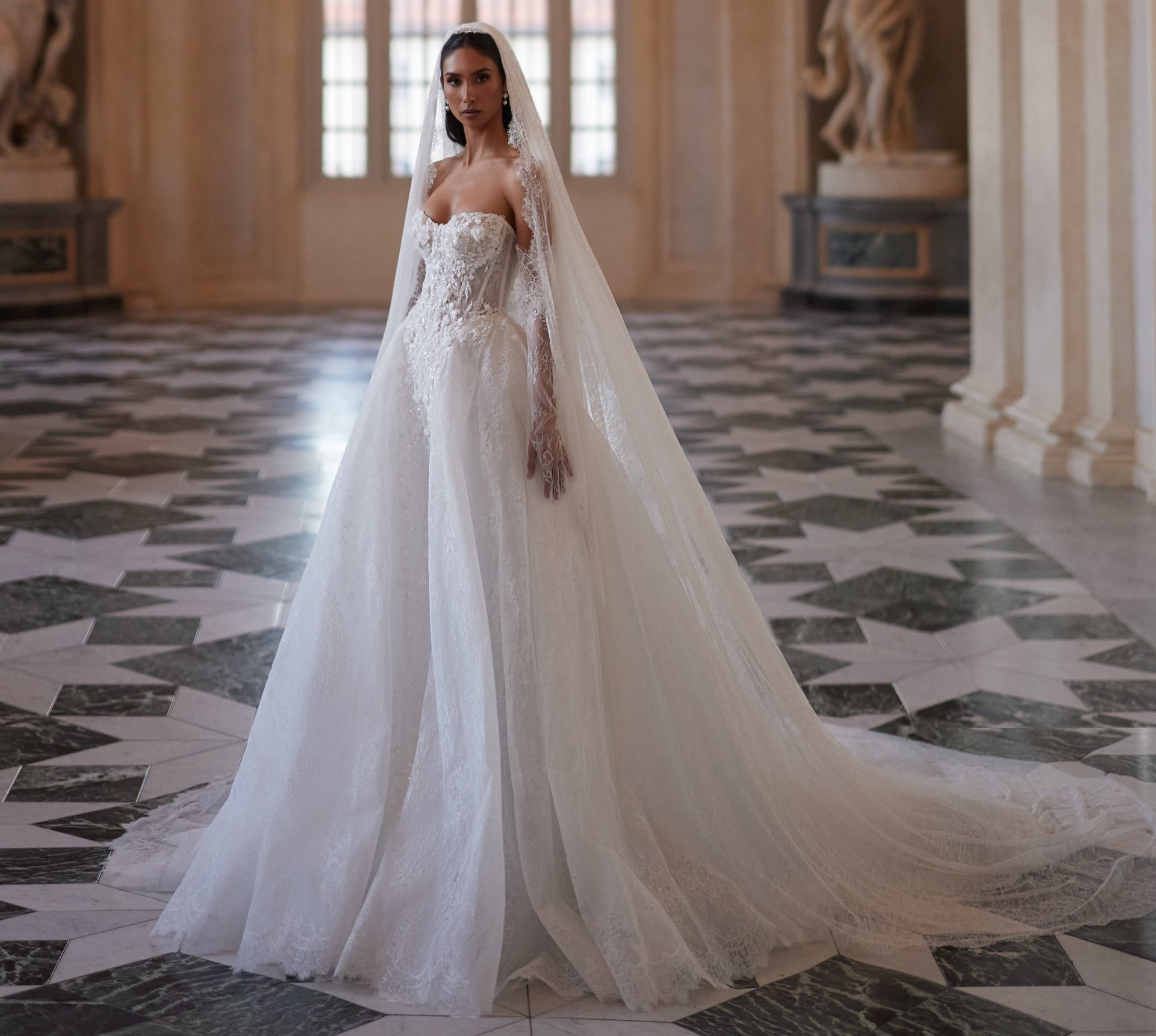 Bride in elegant white wedding gown with long veil and train standing in ornate corridor with geometric floor