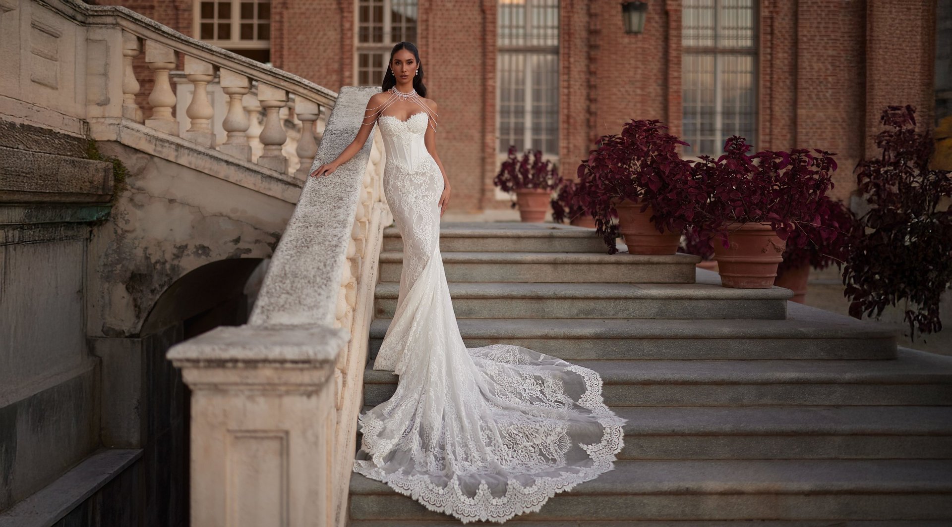 Bride in elegant white lace wedding dress with long train standing on stone steps of historic brick building