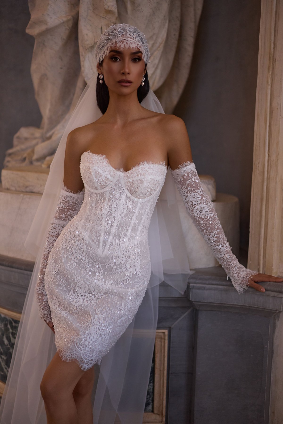 Bride in white wedding gown with veil and train standing in ornate hall with patterned floor and classical sculptures